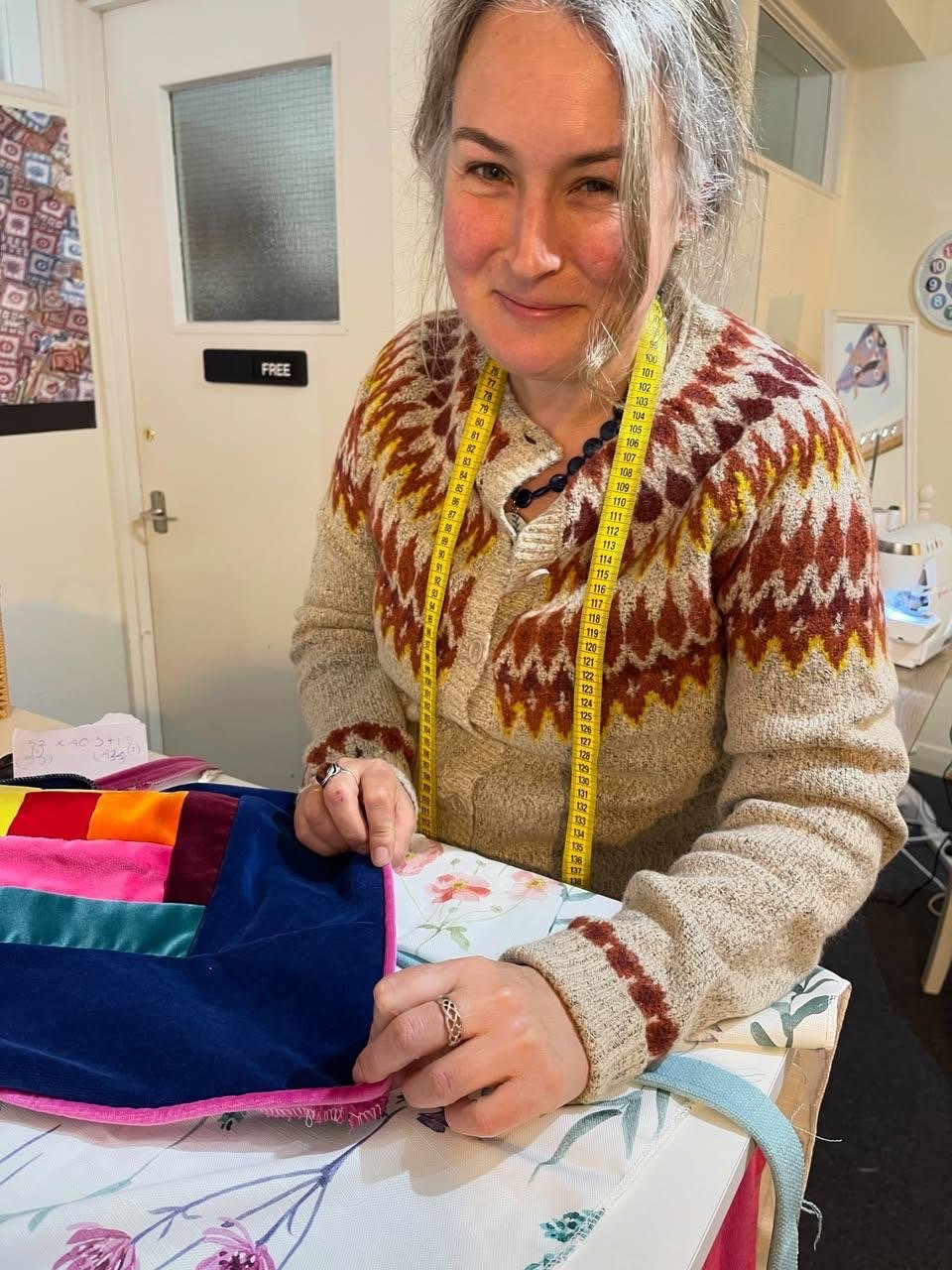 A woman with gray hair and a patterned sweater is holding a colorful fabric bag and looking at the camera in a room with a white door, a clock, and a sewing machine.