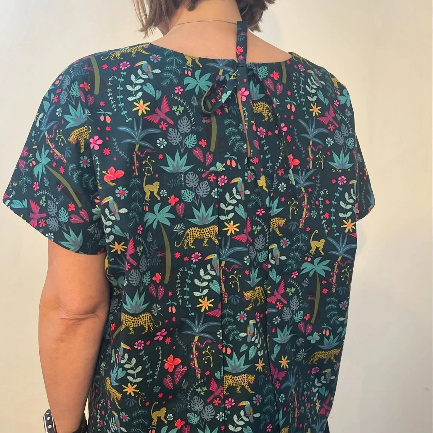 Back of a person wearing a short-sleeved shirt with a colorful jungle pattern featuring various animals and plants, including leopards, toucans, palm trees, and flowers, against a dark background.
