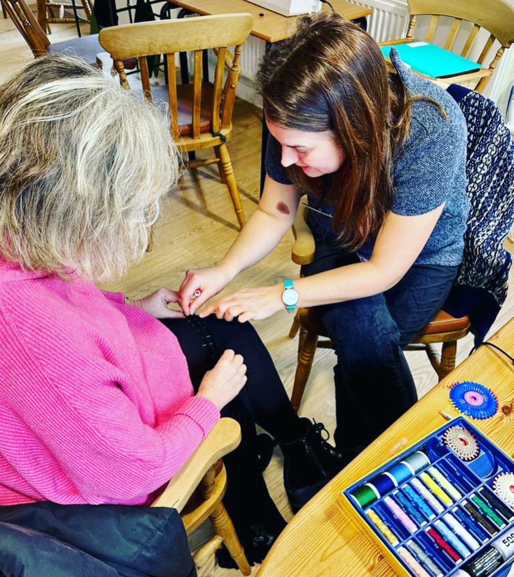 A woman with brown hair, wearing a blue top and blue jeans, is painting the fingernails of an older woman with gray, curly hair, dressed in a pink sweater and black pants, who is sitting in a wooden armchair. The scene takes place in a room with wood
