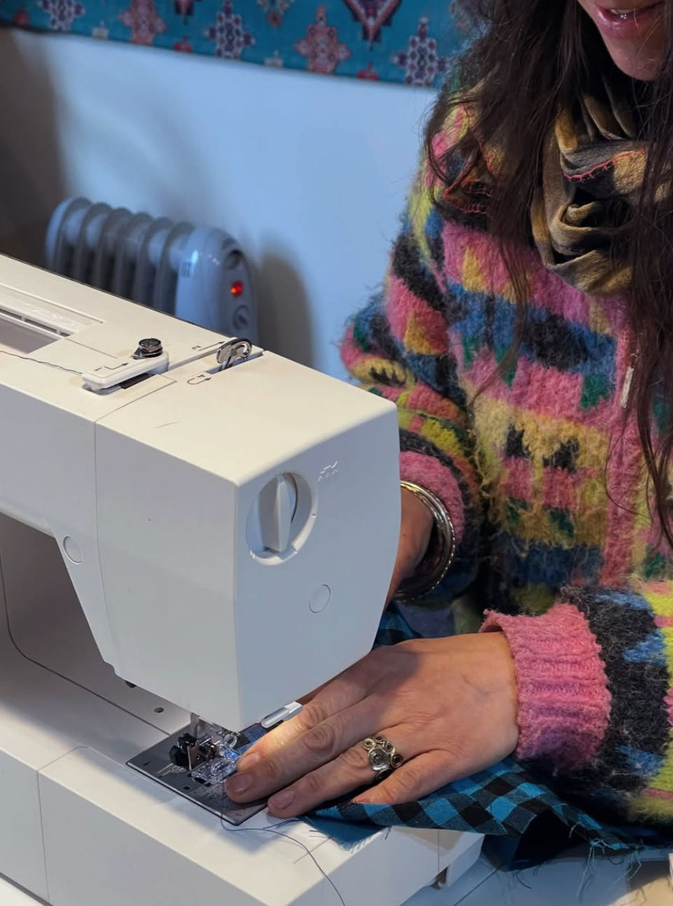 A woman sewing fabric with a white sewing machine, wearing a colorful sweater and a ring on her left hand.