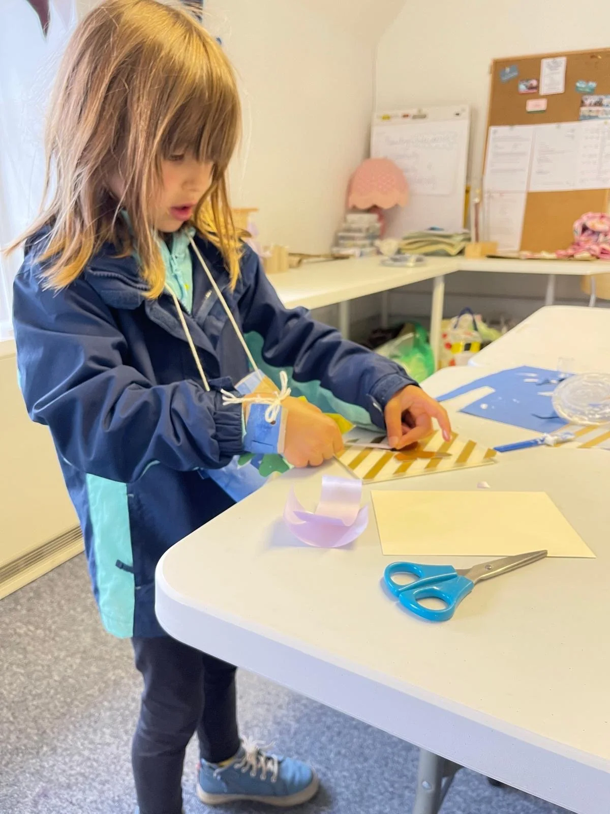 A young girl with light brown hair working on a craft project at a white table. She is wearing a navy blue jacket, black pants, and light blue sneakers. Scissors, paper, and craft materials are on the table, and there is a cork board with papers and 