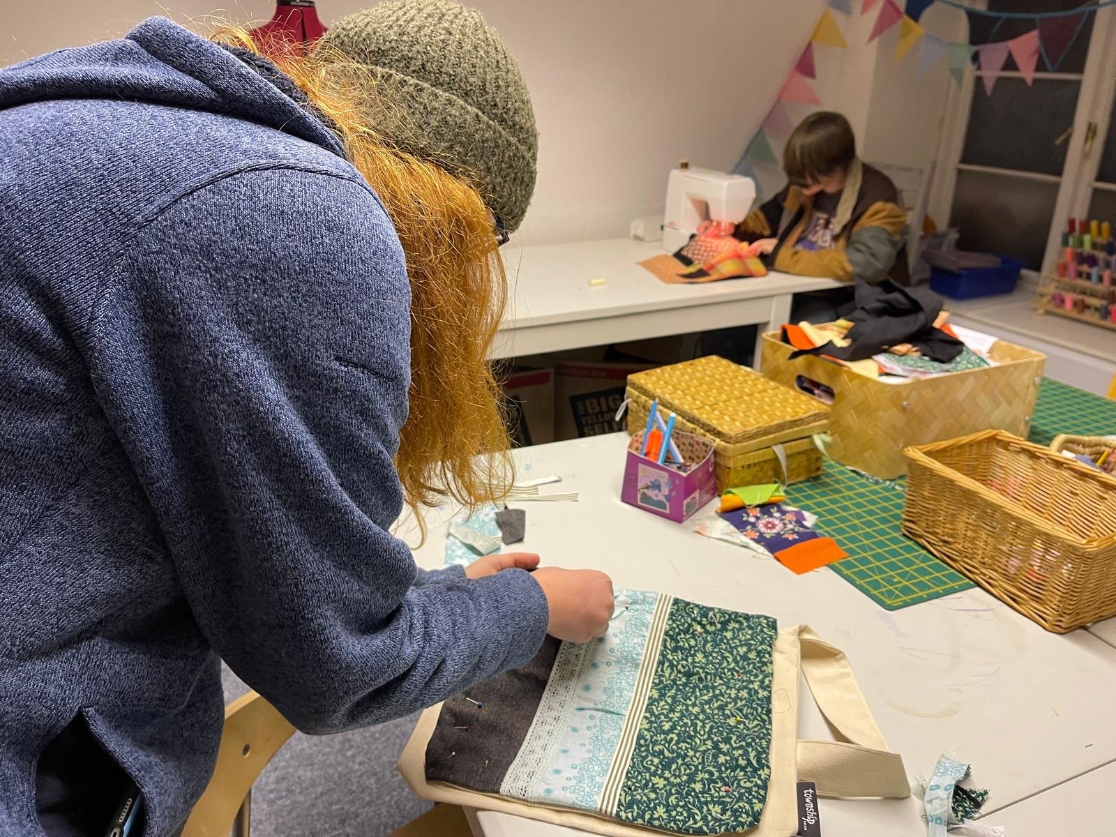 Two people sewing fabric at a craft table, colorful sewing supplies and fabric pieces are scattered on the table. One person is bent over working on a piece of fabric, and another is sitting in the background with sewing materials.
