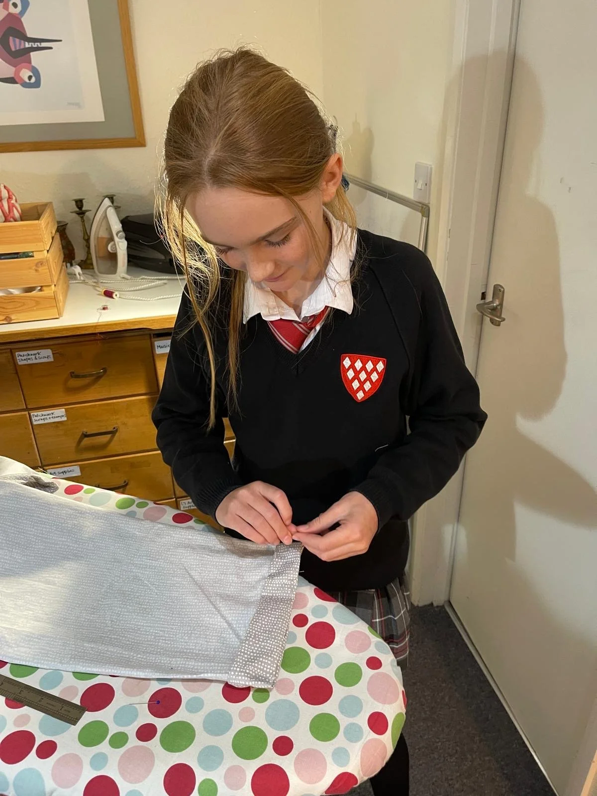 A girl in a school uniform with a crest on her sweater is sewing or stitching a fabric piece on a table covered with a polka dot tablecloth.