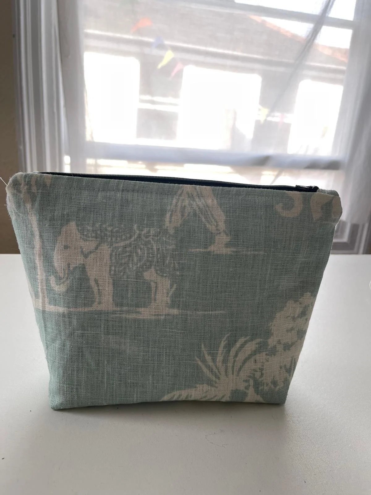 A fabric pouch with an ornate floral and vine pattern, placed on a white surface with a window in the background.