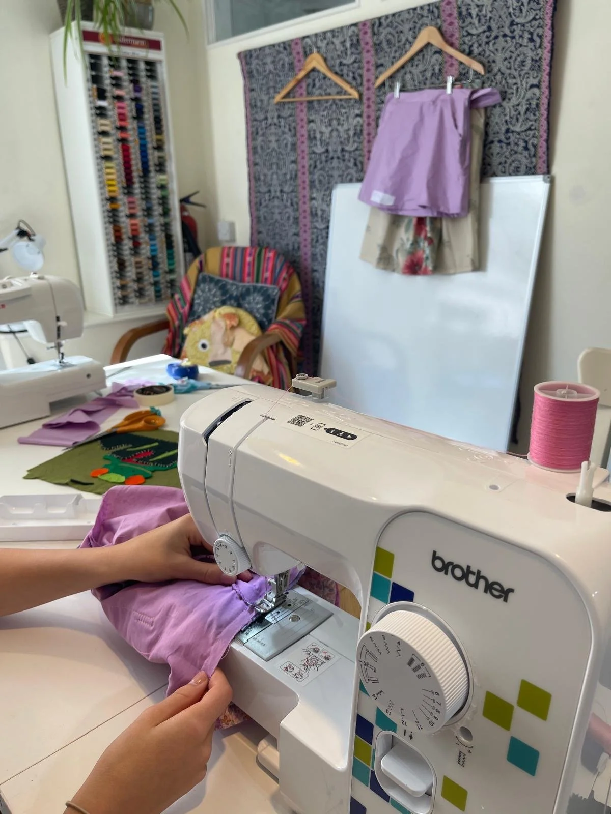 Close-up of hands operating a white Brother sewing machine sewing pink fabric, with a sewing workspace in the background including colorful thread spools, fabric, and sewing supplies.