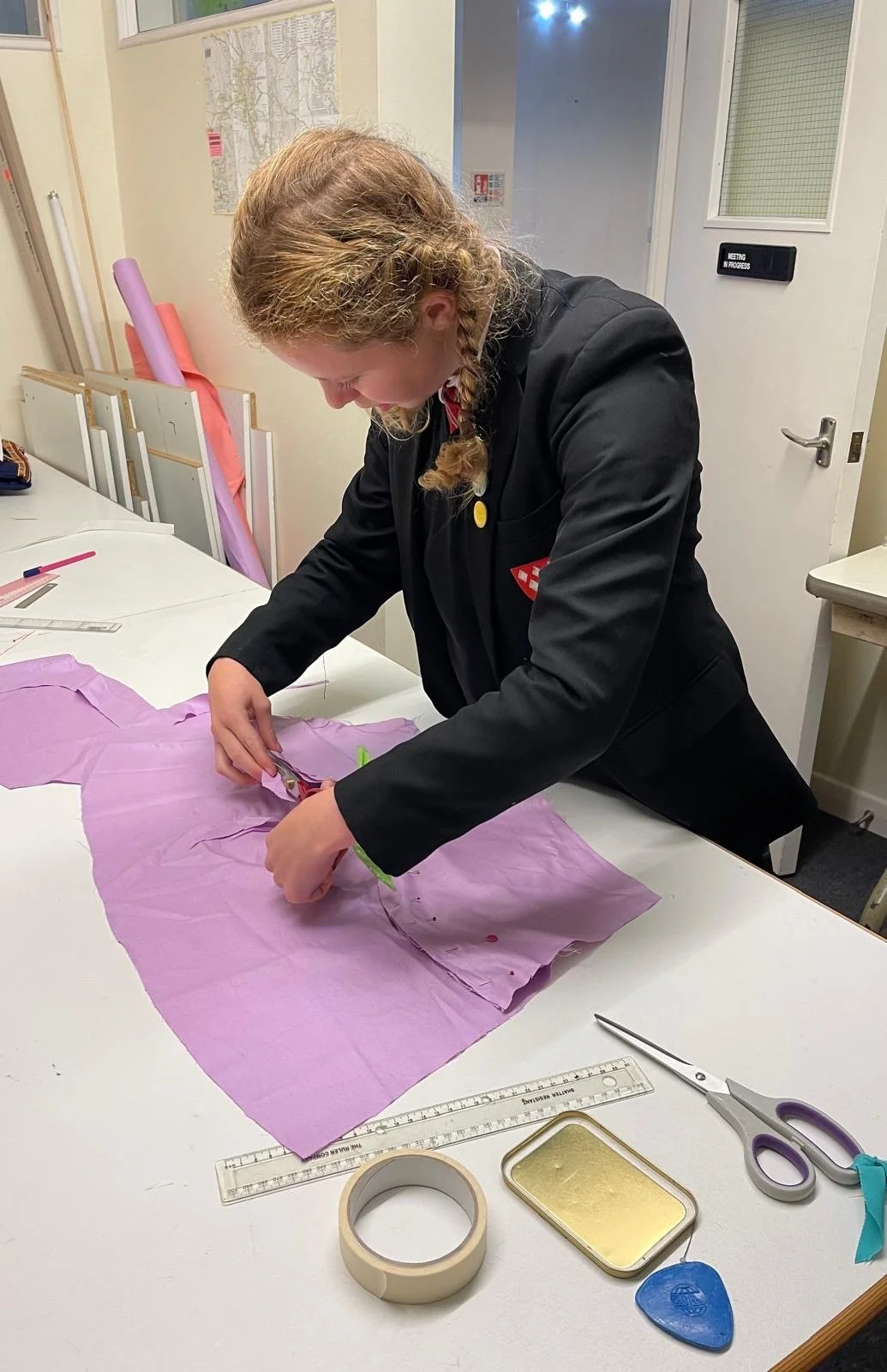A woman sewing on a pink piece of fabric on a white table, using scissors. There are various sewing tools and supplies on the table, including rulers, tape, a pin cushion, and fabric.