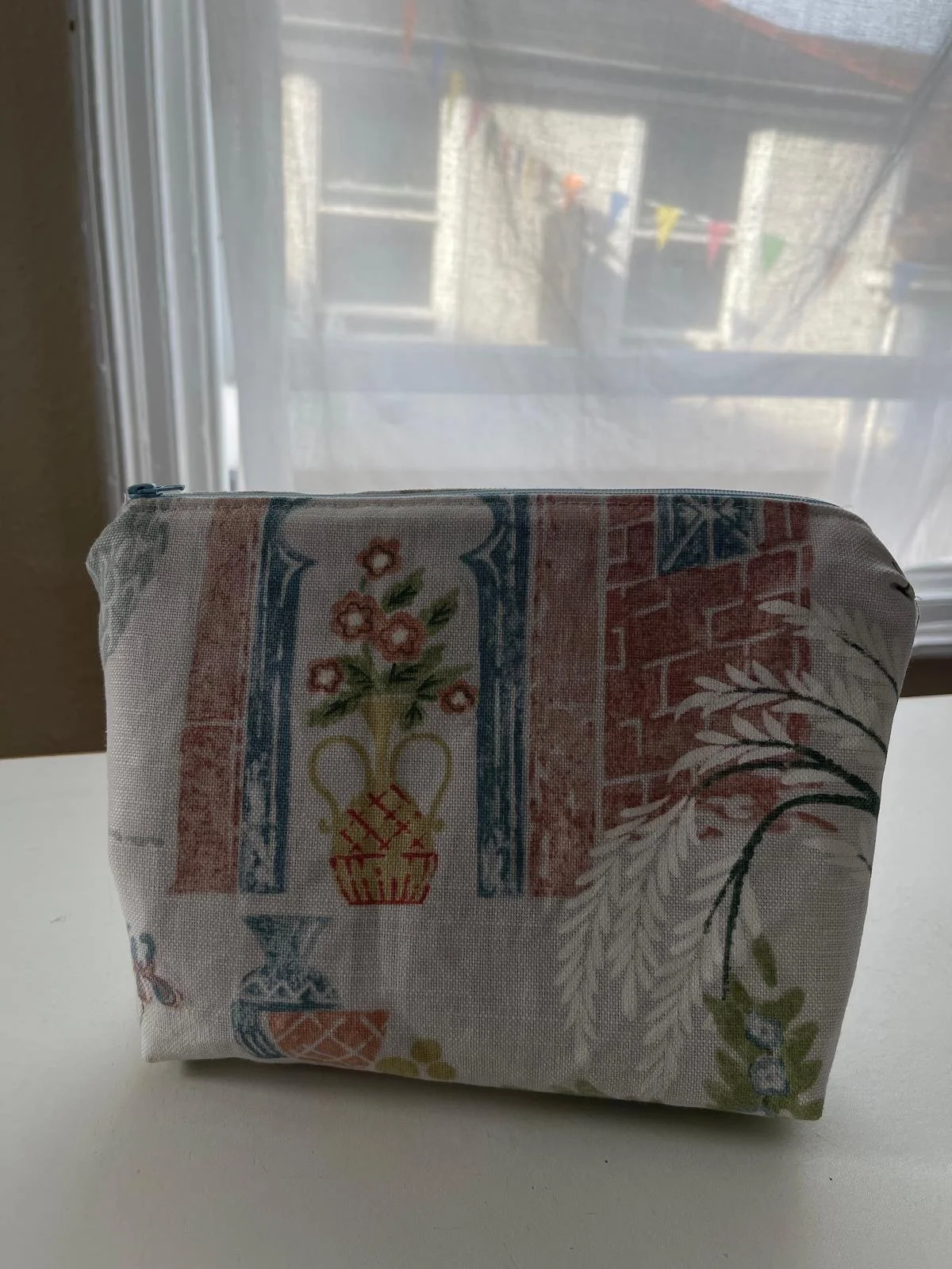 A patterned fabric pouch with floral and decorative motifs, placed on a white surface near a window with a sheer curtain. The window shows a blurry exterior view with bunting decorations.