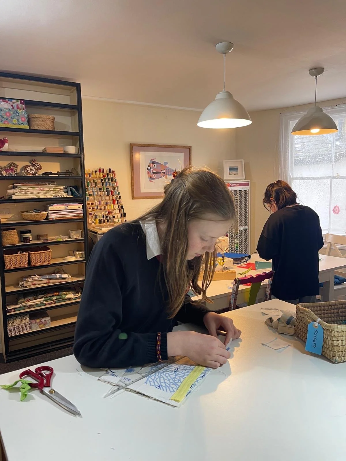 Two women working at a craft table in a well-lit room with sewing and craft supplies, including scissors, fabric, and thread, in the background.