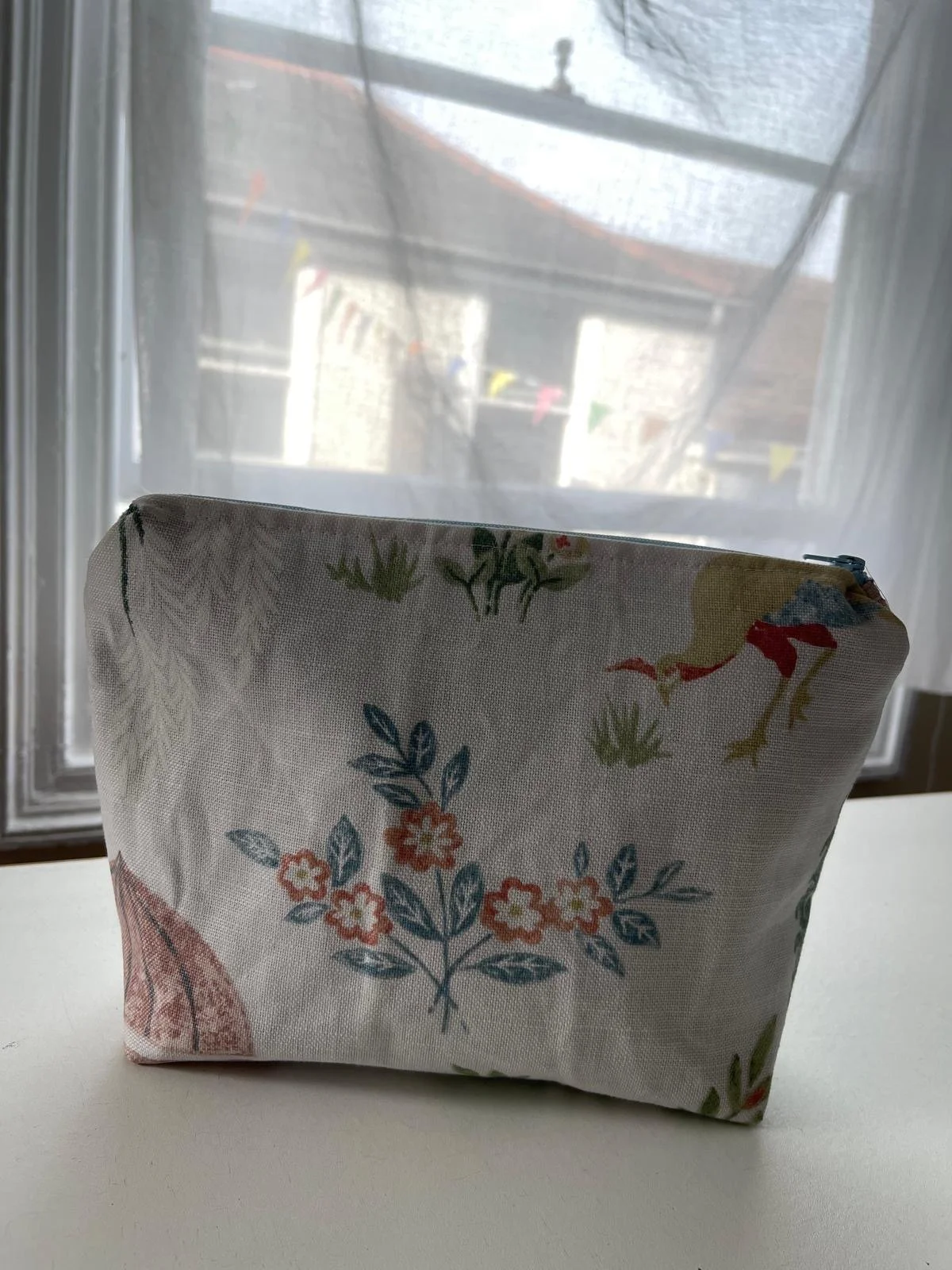 A cloth bag with a floral and bird pattern sitting on a white surface in front of a window with sheer curtains, through which a building and colorful bunting are visible outside.