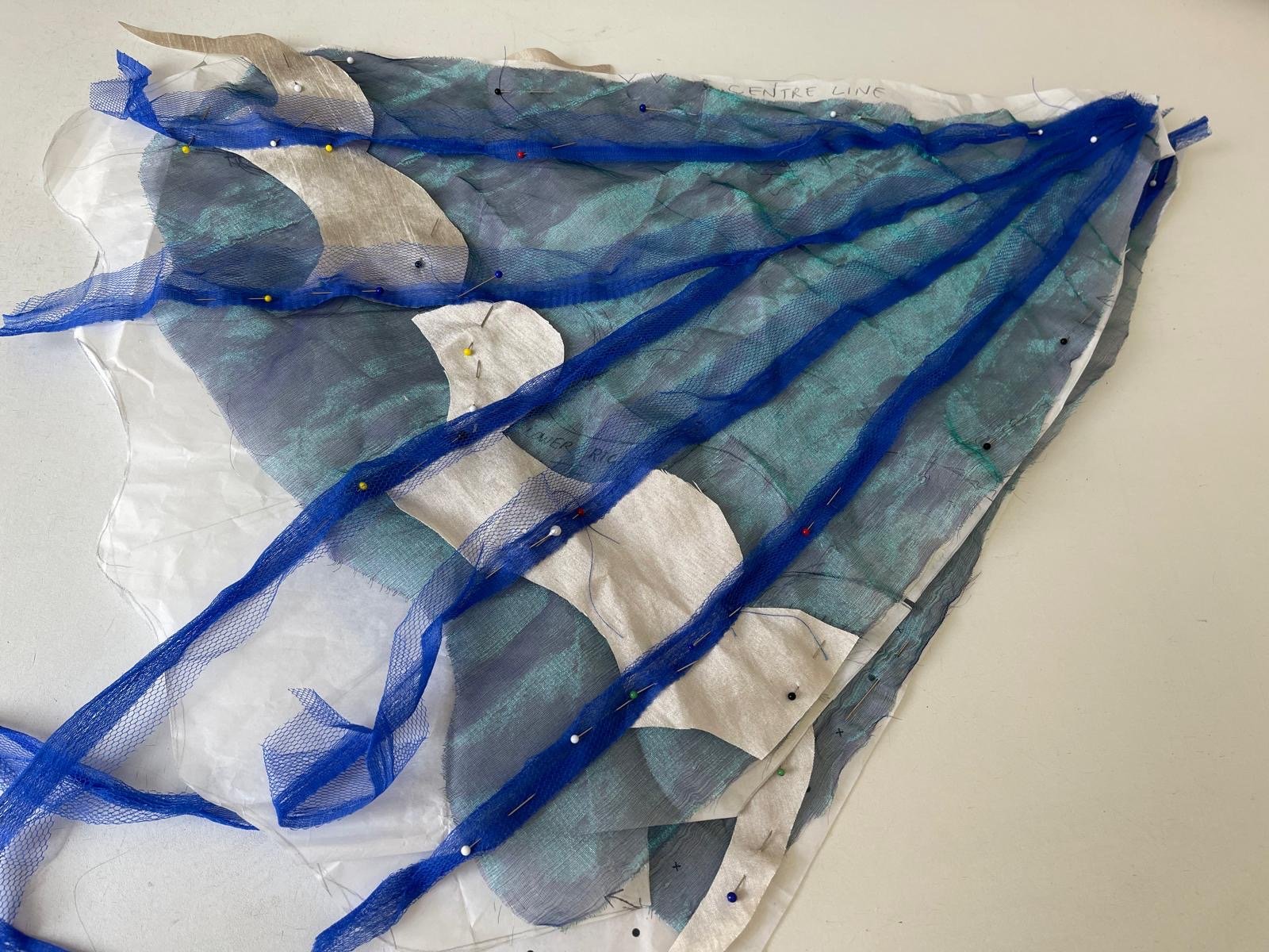 A layered fabric art piece with blue, silver, and gray fabrics, assembled with pins and sewing threads, displayed on a flat surface.