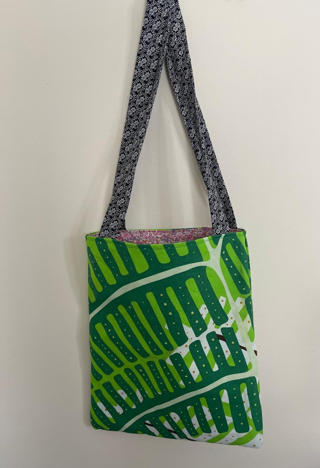 A tote bag with a green and white leaf pattern hanging on a white wall by black and white patterned handles.
