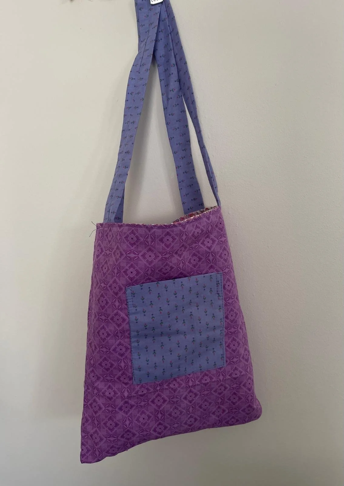 A purple quilted tote bag with a decorative square pocket on a plain wall.