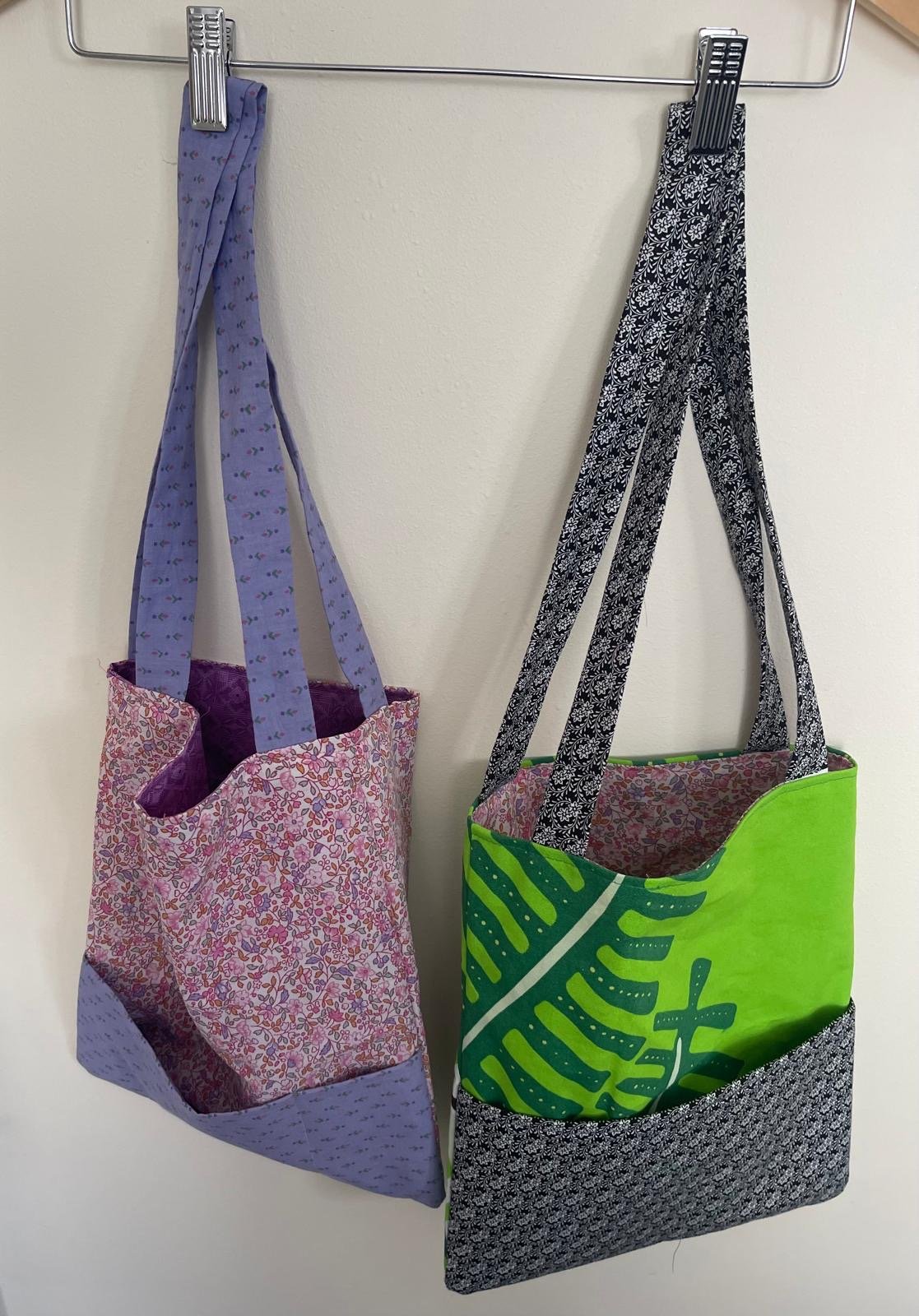 Two fabric tote bags hanging on hooks from a metal rack, one with a purple and pink floral pattern, the other with black and white patterns and a green fabric section.