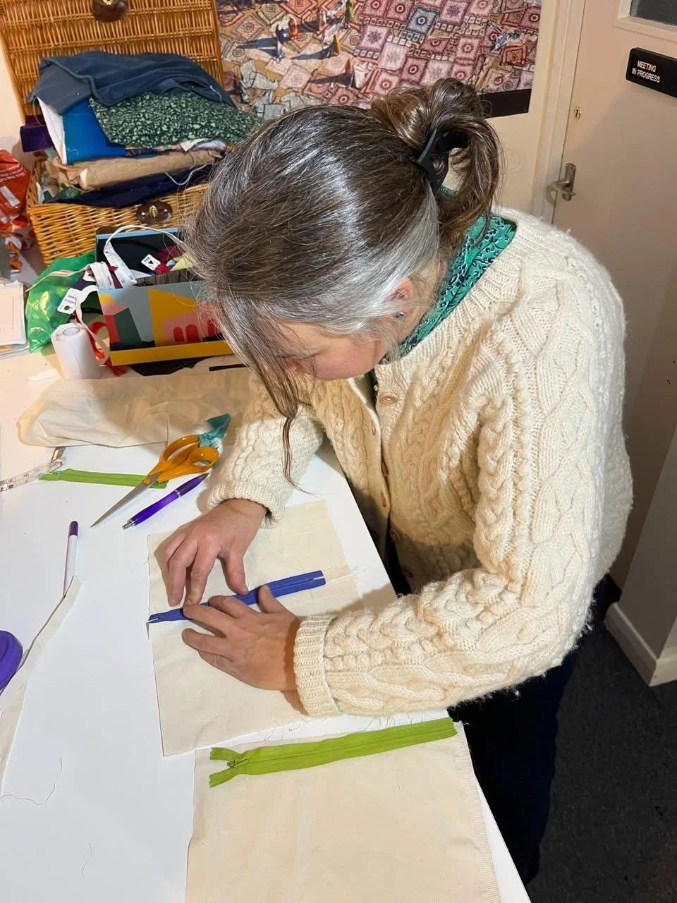 A woman with gray hair tied back in a ponytail, wearing a cream-colored cable knit sweater and teal bandana, is sewing or working on a craft project at a white table. The table has fabric, scissors, pens, and zippers. In the background, there are sta