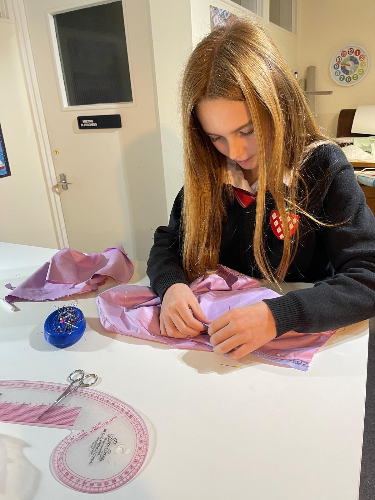 A young girl with long red hair sewing pink fabric at a sewing table. There are sewing tools and a pink plastic measuring guide on the table, with a pin cushion nearby. She is wearing a black sweater and a school uniform with a red and white emblem o