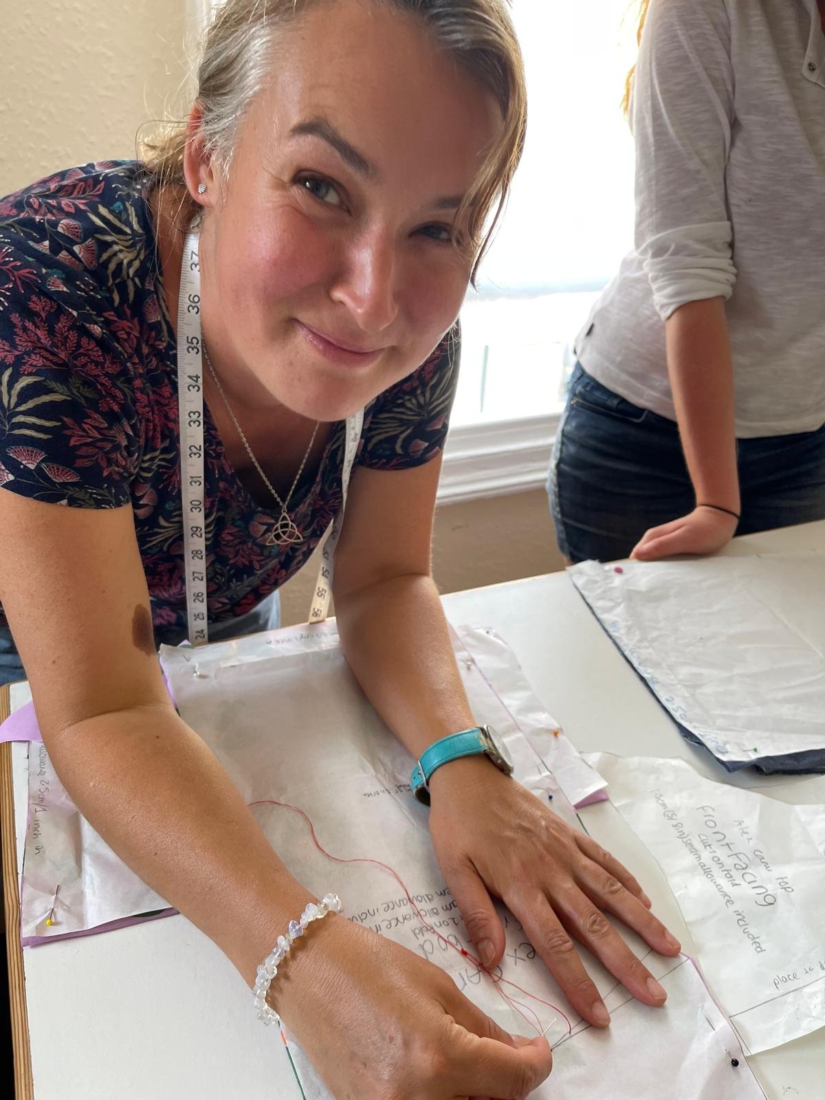 A woman leaning over a table, smiling at the camera, with a measuring tape around her neck, working on a project with papers and drawings on the table, and another person standing beside her.