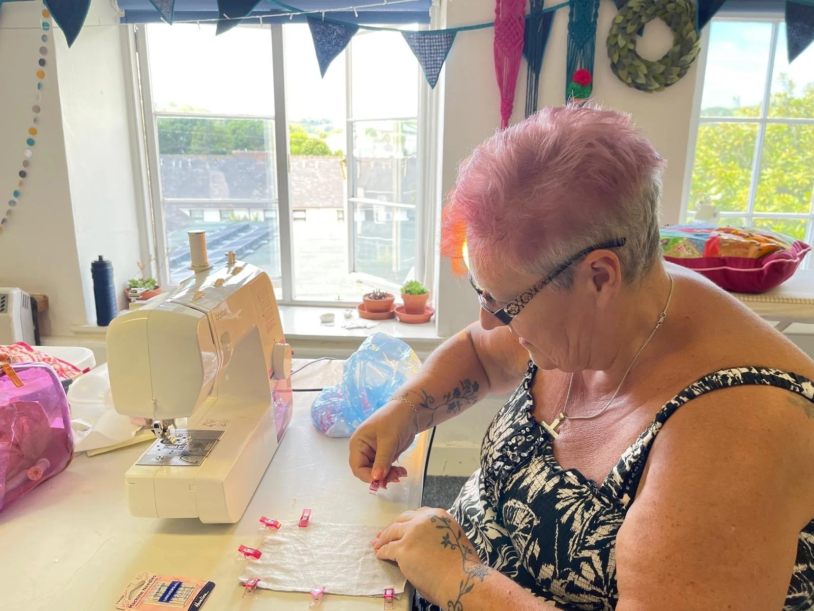 An older woman with short pink hair and glasses, wearing a black and white patterned tank top, is sitting at a table sewing or working with small fabric pieces. She is indoors, near a large window with a view of a backyard and blue sky. The table has