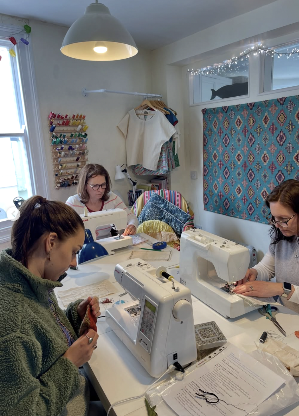 Three women working on sewing machines in a bright, colorful sewing room with spools of thread on the wall, hanging clothes, and decorative textiles. A window with fairy lights and a fabric wall hanging are also visible.