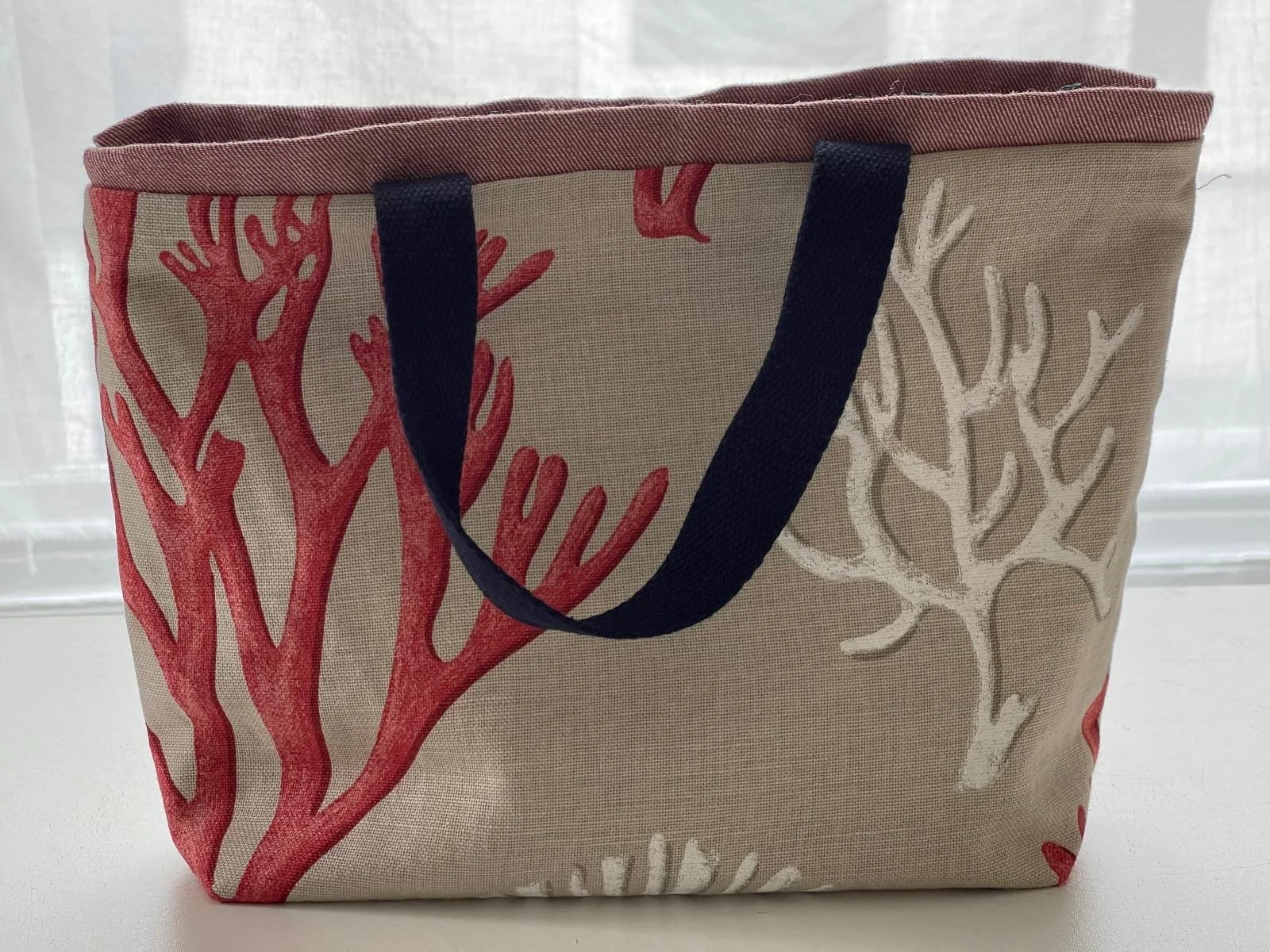 A tote bag with a beige background decorated with red and white coral reef designs, black handles, and a red top edge.