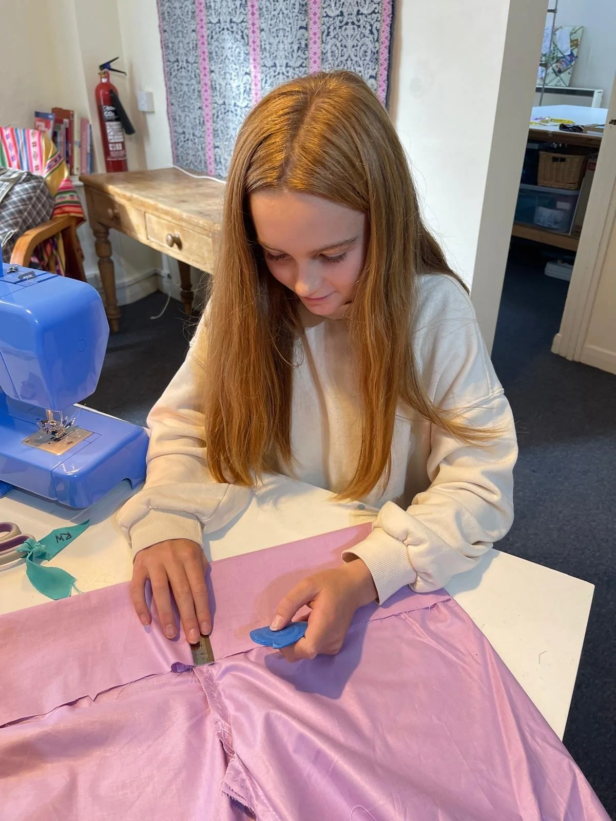 A young woman with long red hair, wearing a cream-colored sweatshirt, sewing pink fabric on a white table in a room with sewing supplies and a sewing machine.