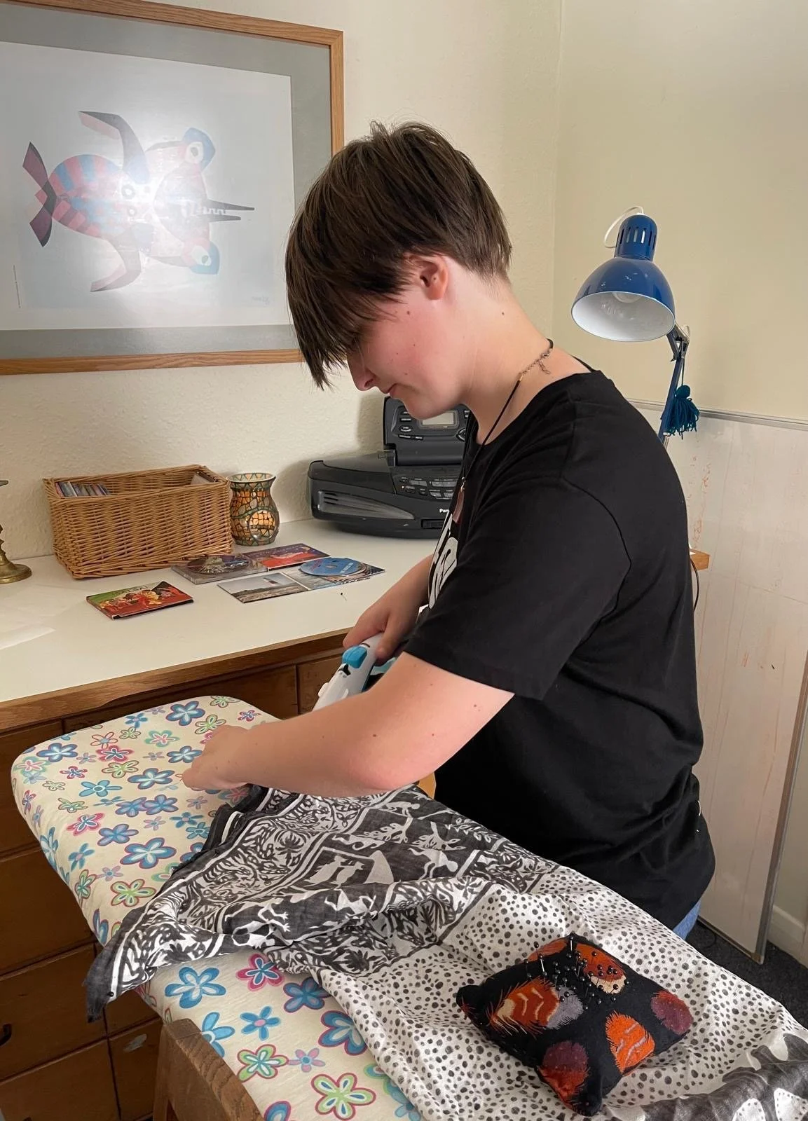 A person sewing fabric on an ironing board in a room with a framed colorful abstract artwork, a wicker basket with cards, and a small lamp on a desk.