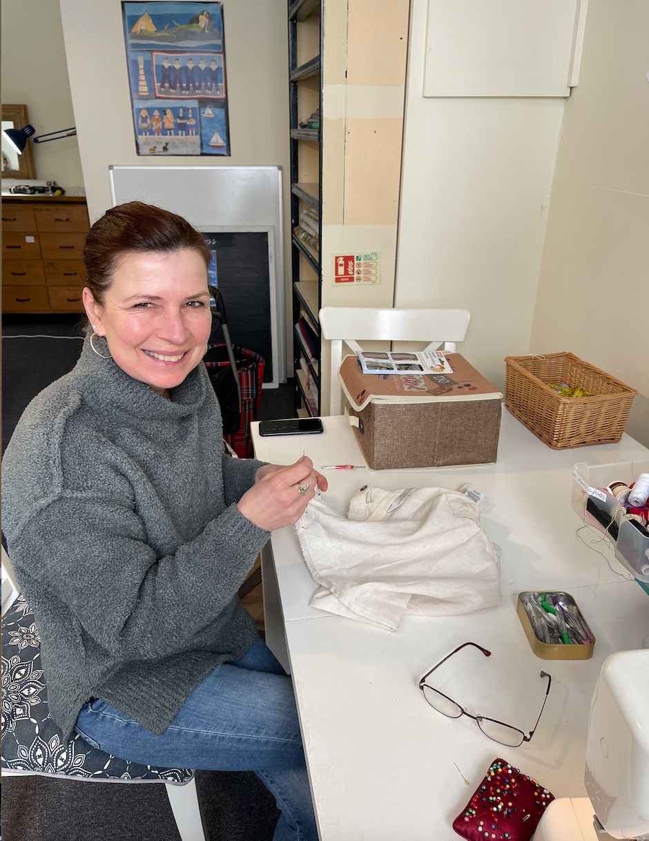 Woman sewing or crafting at a table, smiling, with sewing supplies and glasses nearby in a room with shelves and artwork.