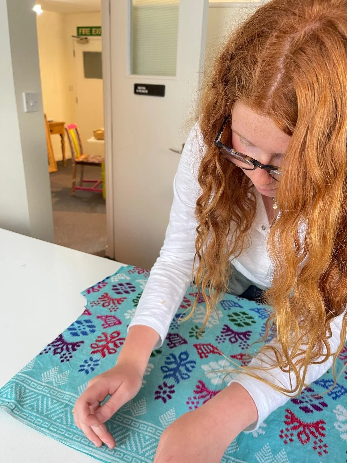 A woman with long red hair and glasses is folding a colorful, patterned tablecloth on a white table in a room.