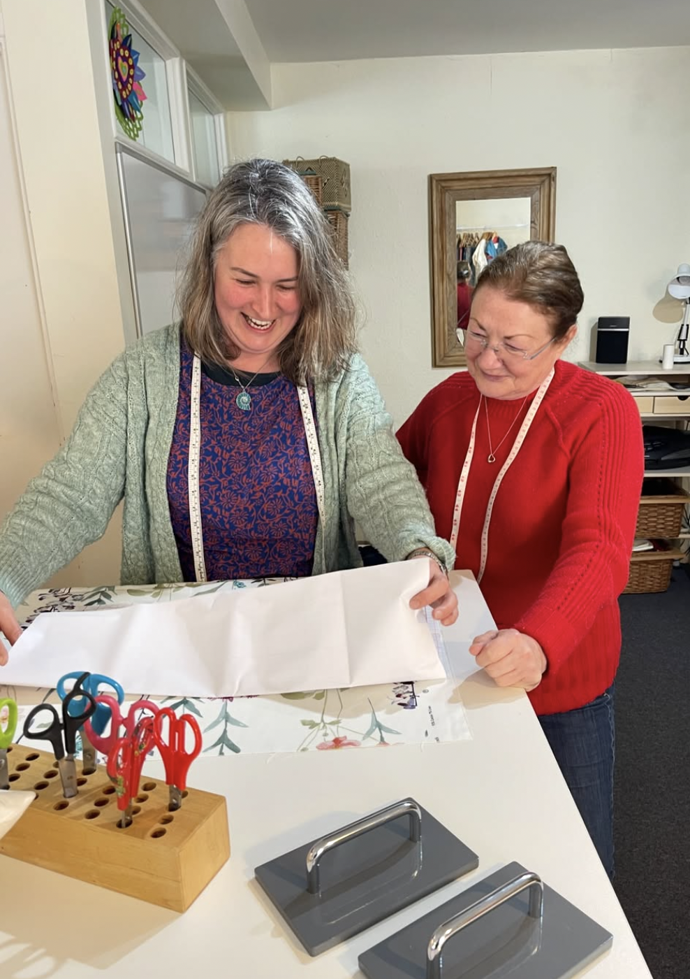 Two women in a craft room unwrapping a large wrapped gift, with scissors and paper on the table.