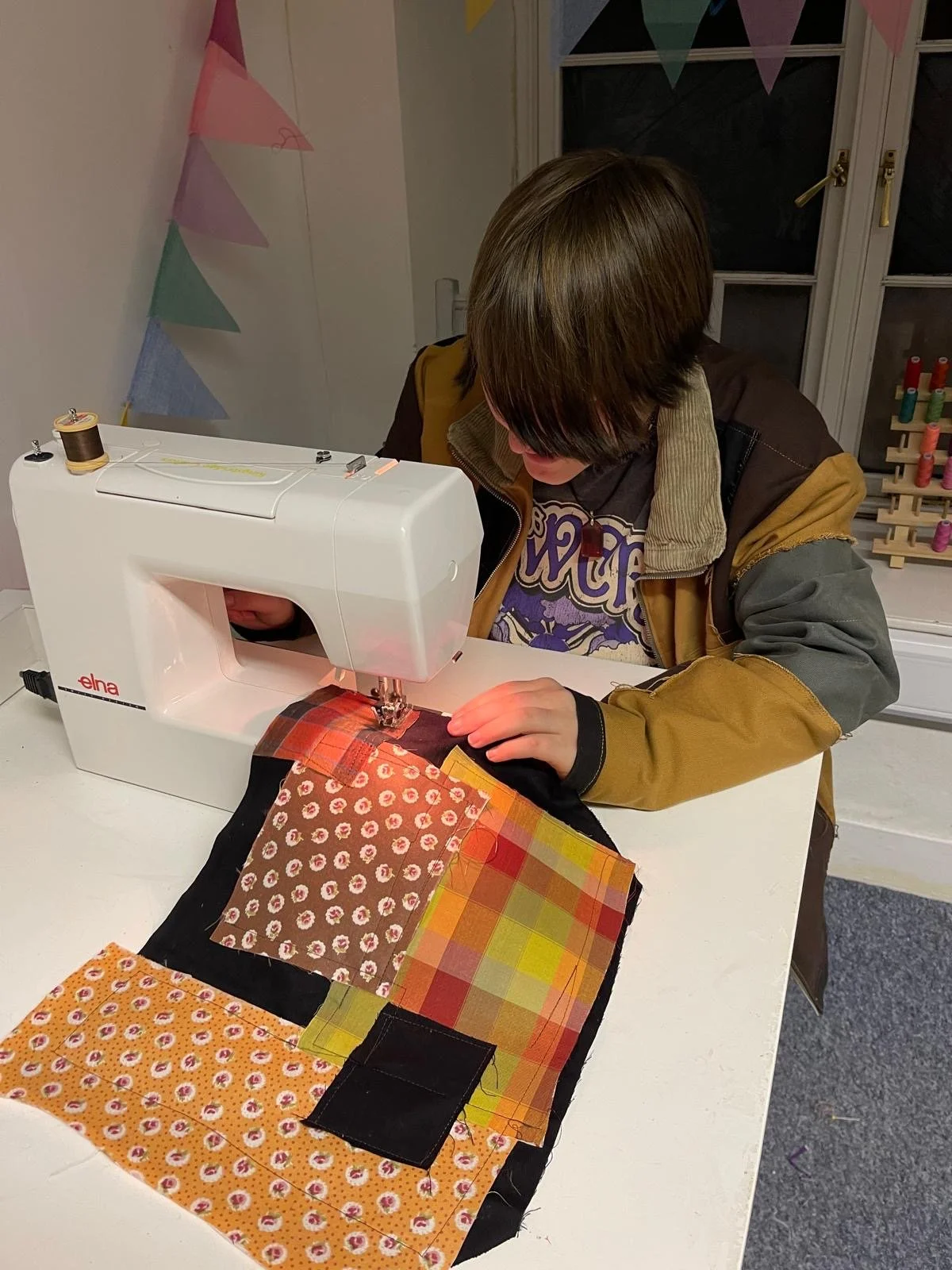 A person sewing colorful fabric squares on a white sewing machine in a room decorated with pastel flags.
