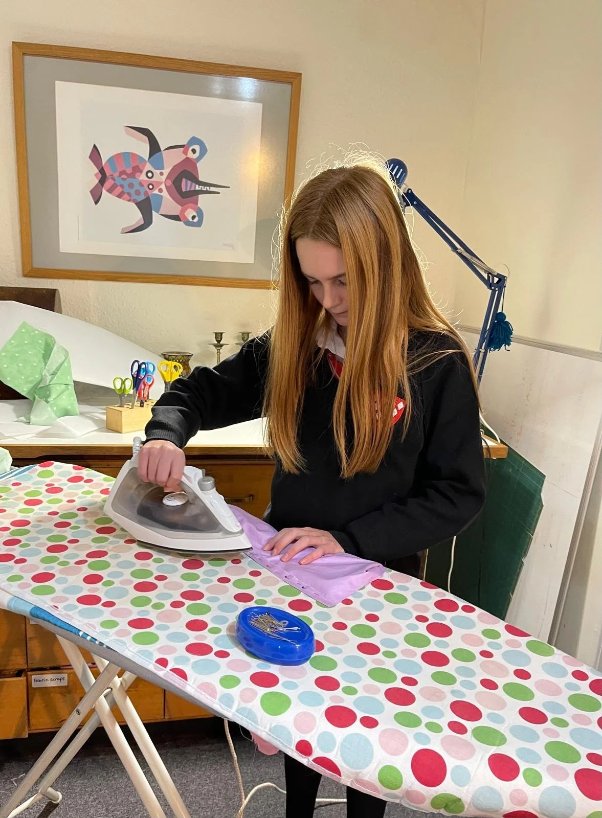 A young woman with long red hair ironing purple fabric on a colorful polka dot tablecloth.
