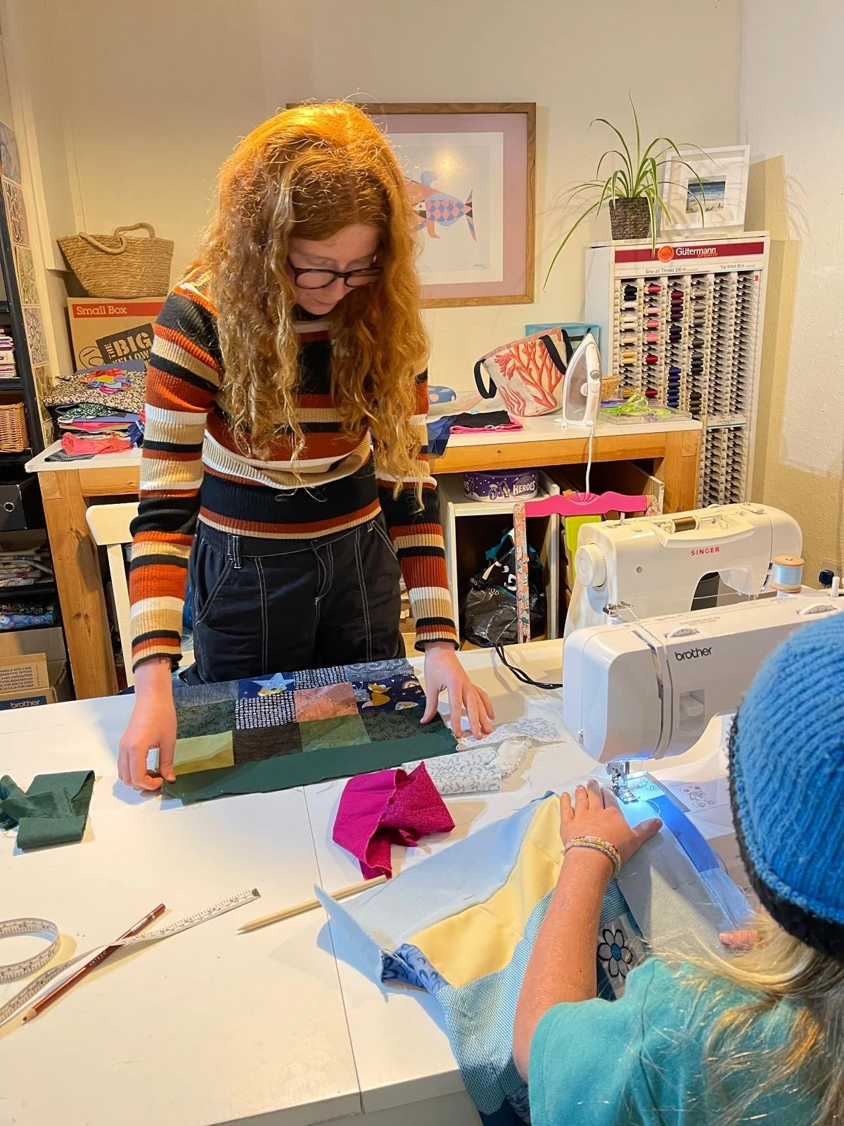 Two women are sewing fabric pieces on a white table in a craft room. One woman, with long curly hair and glasses, is standing and looking down at the fabric, while the other, with long blonde hair and a blue hat, is sitting at a sewing machine. The t