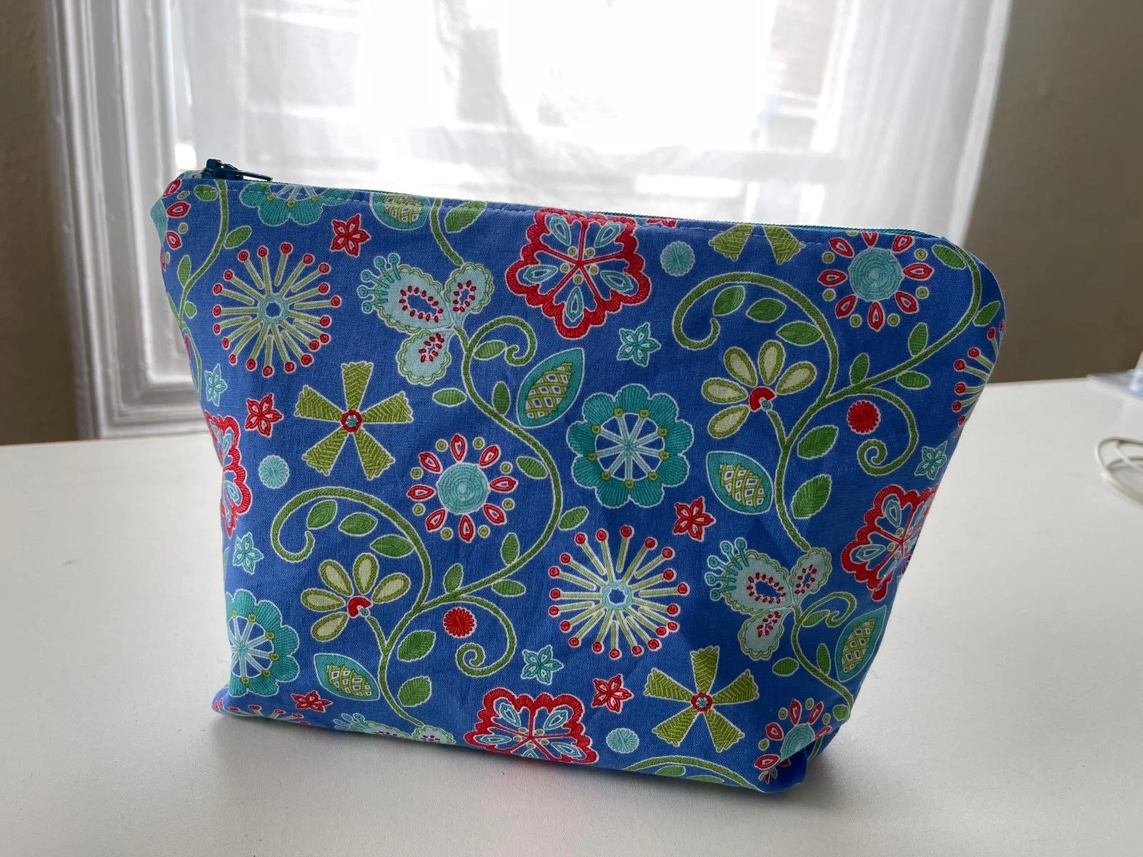 A colorful cosmetic or toiletry bag with a floral pattern on a white surface, with a window in the background.