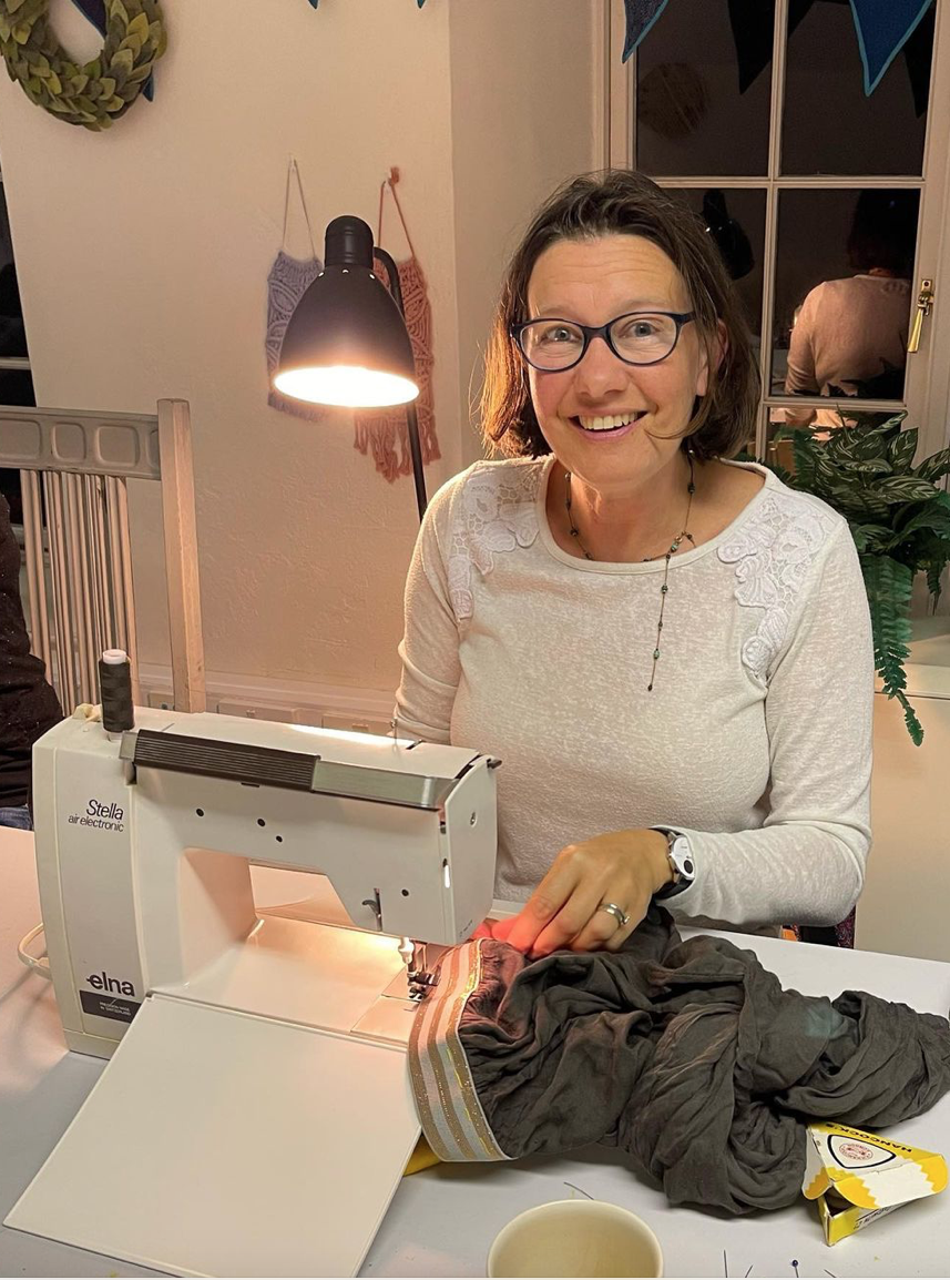 A woman with glasses, smiling, sewing fabric on an electronic sewing machine at a table. The scene is indoors with a lamp, wall decorations, a window, and a potted plant in the background.