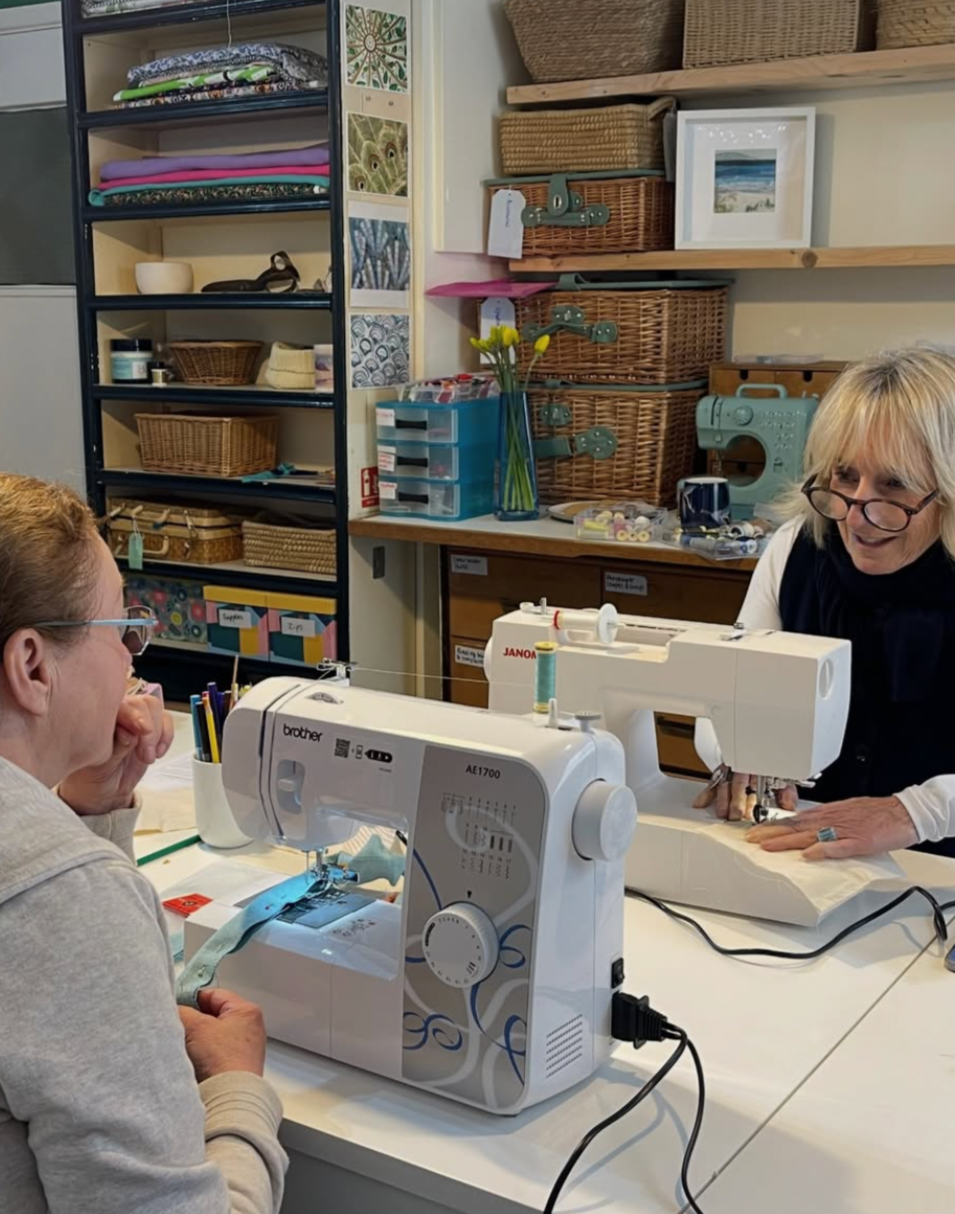 Two women sewing together in a craft room, using sewing machines, surrounded by shelves with fabric, baskets, and craft supplies.