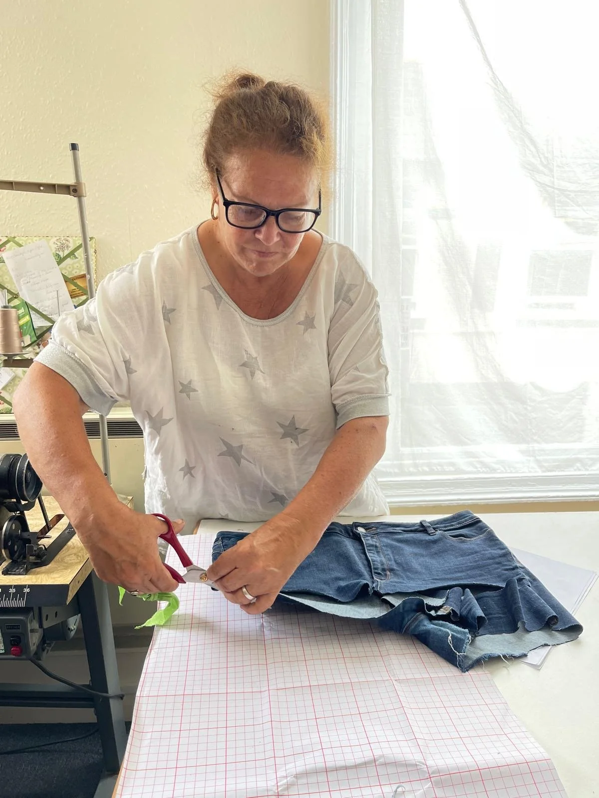 A woman cutting fabric with scissors on a table, with folded denim jeans nearby, in a bright room with a large window.