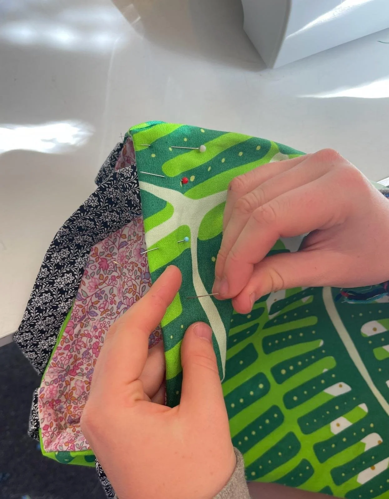 Person sewing a quilt or fabric piece with colorful printed fabric using straight pins on a white surface.