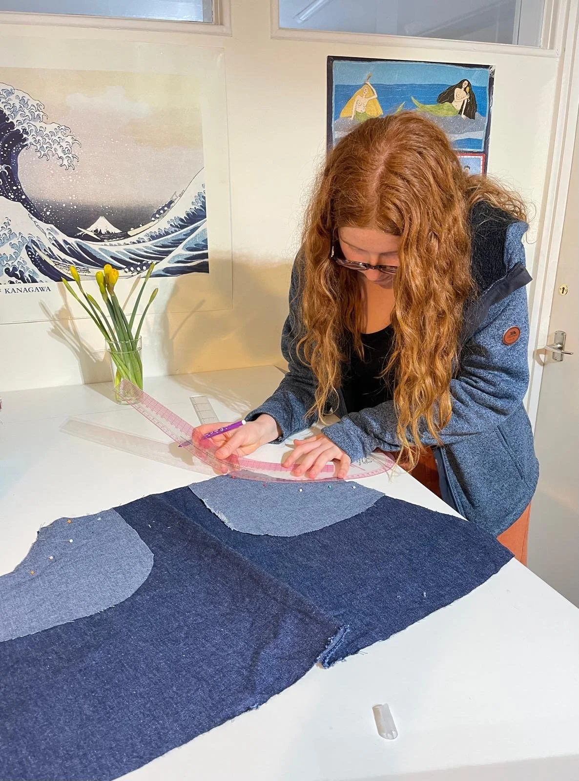 A woman with long curly red hair measuring a piece of dark blue denim fabric with a protractor and ruler on a white table. There is a vase with yellow flowers, a ruler, and a picture of a wave and volcano in the background.