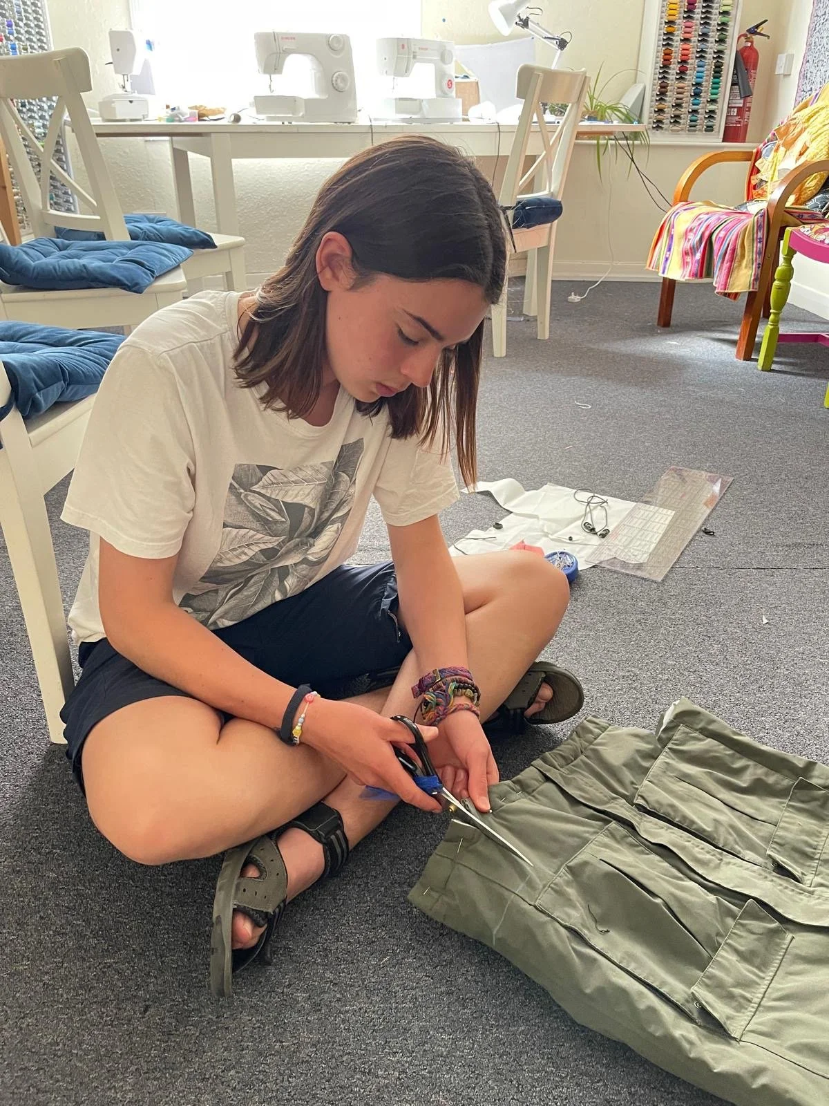 A young girl sitting on the floor, cutting fabric with scissors. She is surrounded by sewing and crafting supplies, with sewing machines on the desk behind her and a color thread rack on the wall.