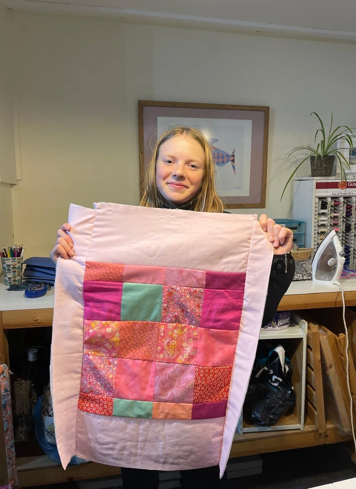 A young girl holding a pink quilt with a patchwork design, standing in a sewing room.