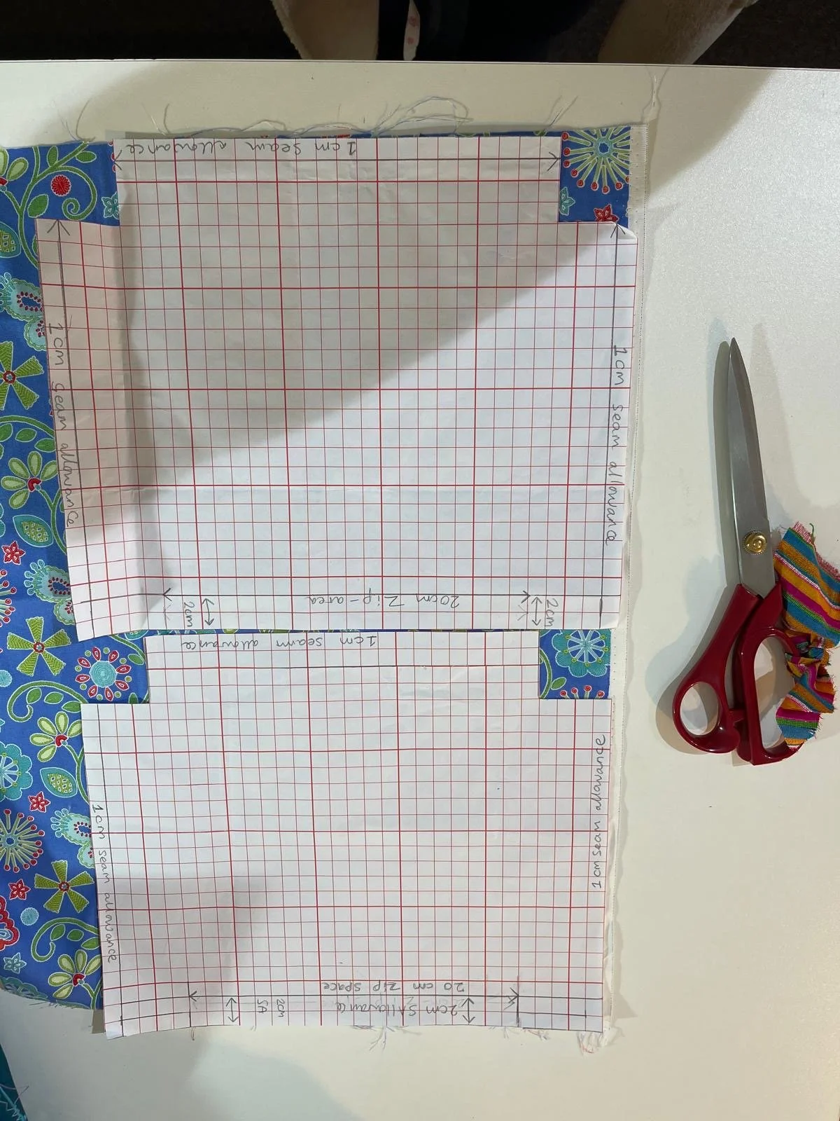 Two sheets of graph paper with measurements and notes, taped to a table with colorful patterned tape. Red scissors and multicolored striped ribbon are on the right side of the image.