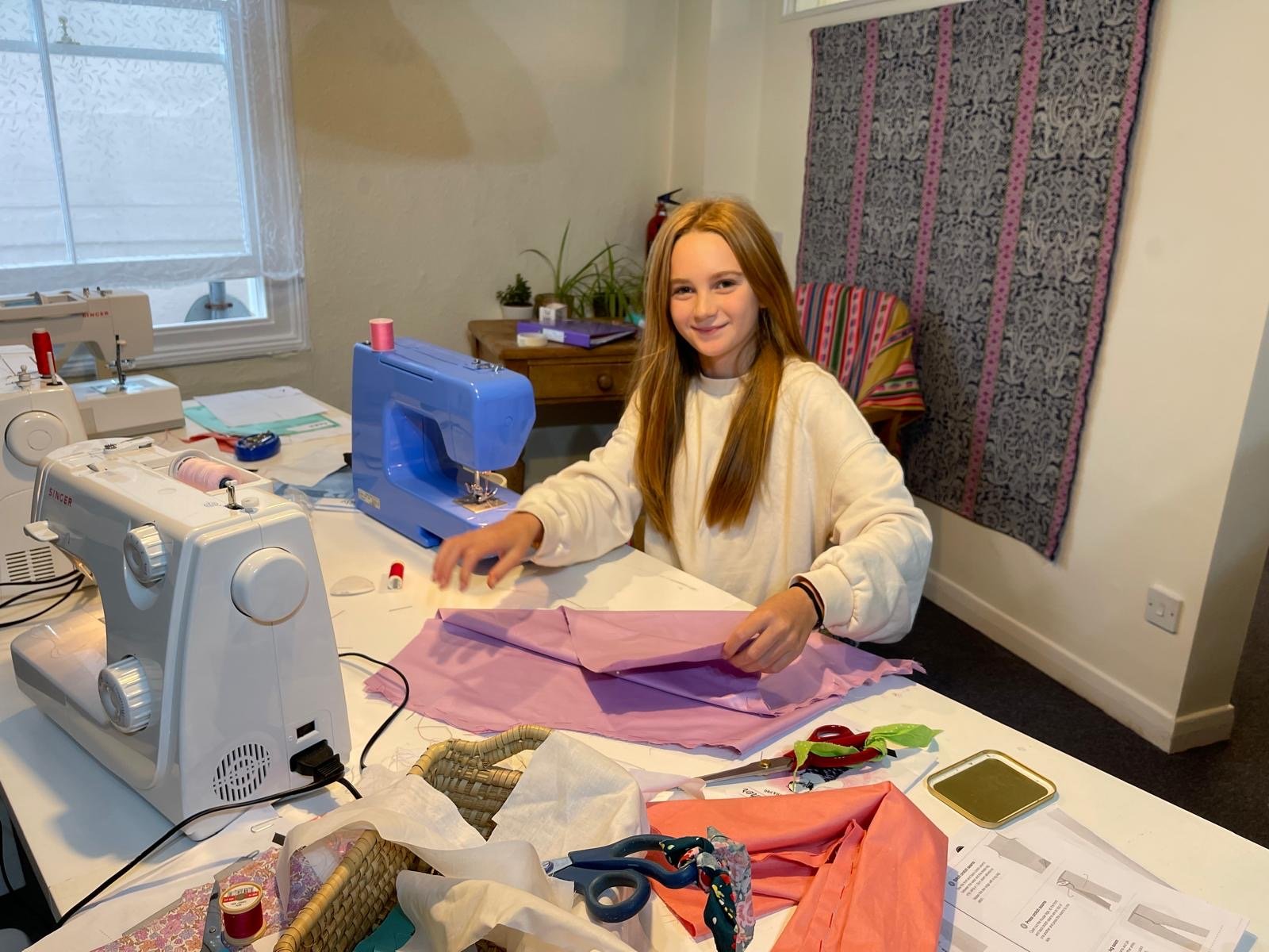 A young girl with long red hair sitting at a sewing table with sewing machines, fabric, scissors, and sewing supplies, working on a pink piece of fabric.