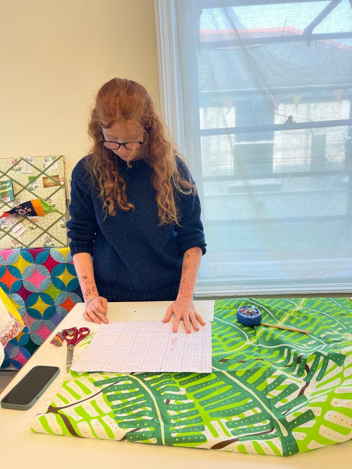 A woman with long red hair and glasses stands at a table, looking down at a grid paper. She wears a dark sweater and has henna designs on her arms. The table has scissors, a smartphone, and a small round container, and there is a green leafy patterne