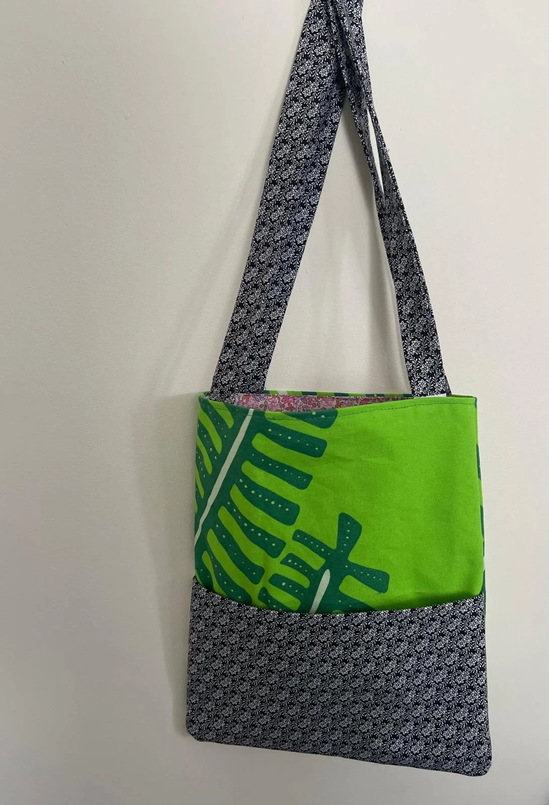 A tote bag hanging on a wall, with black and white patterned straps and a body made of bright green fabric featuring a large palm leaf design.