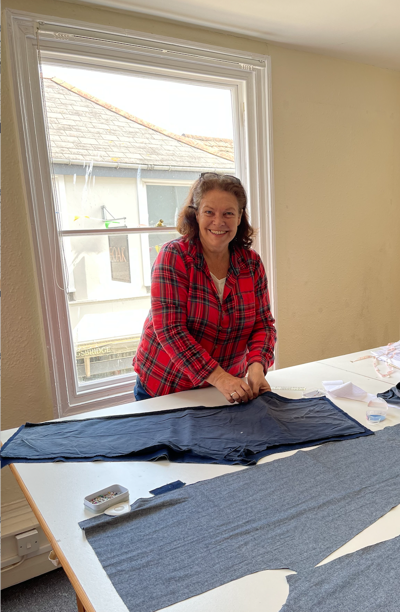 A woman standing at a table covered with fabric, smiling, inside a room with a large window and a house visible outside.