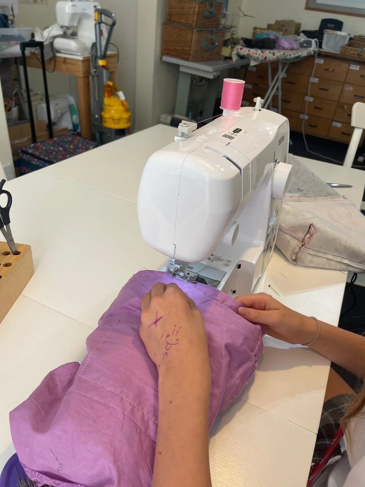 A person sewing fabric on a white sewing machine in a craft room. The person's hand is on purple fabric, with markings on their hand.