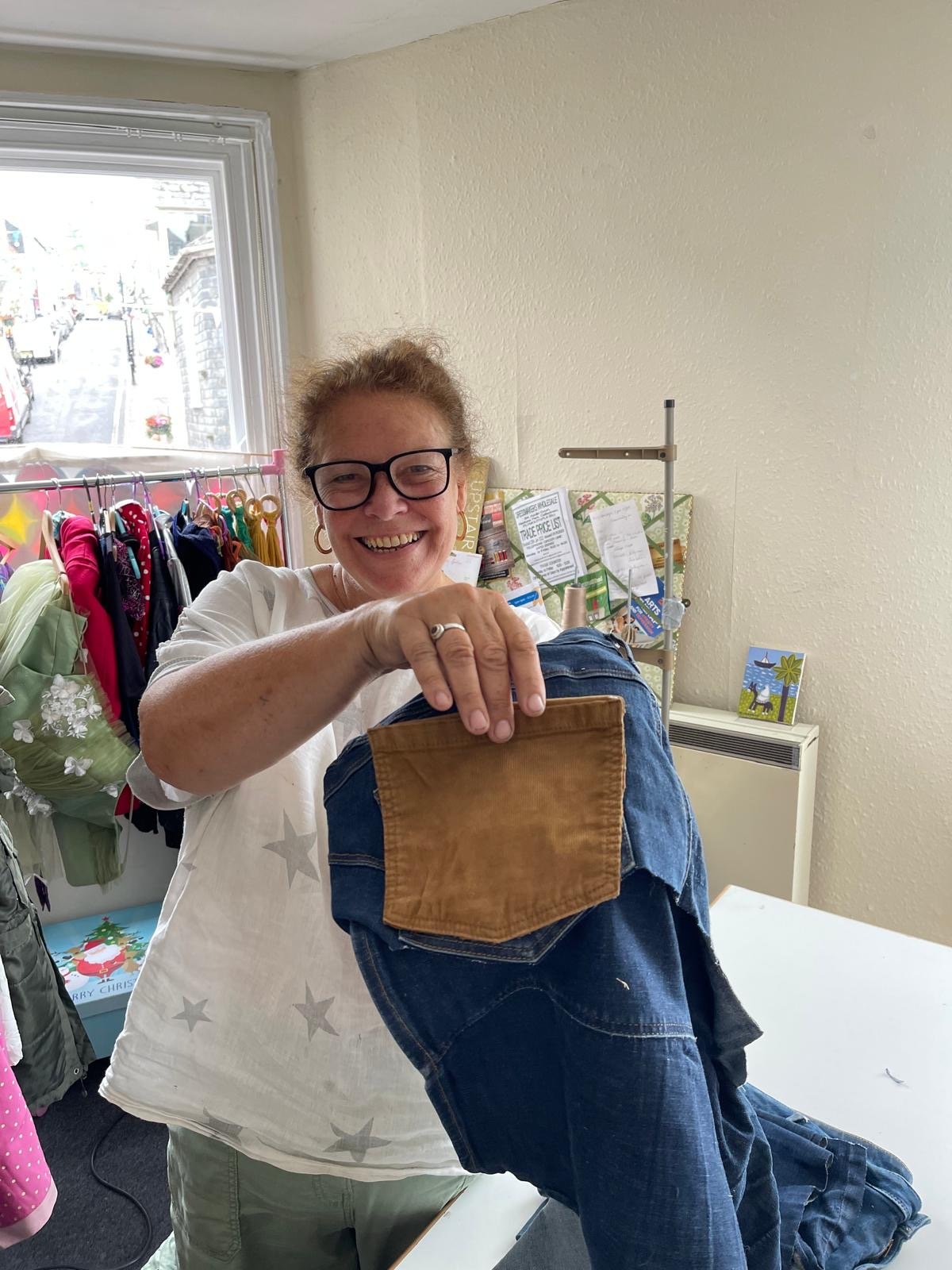 A smiling woman with glasses holding a denim and leather bag while standing behind a person wearing jeans inside a clothing store.
