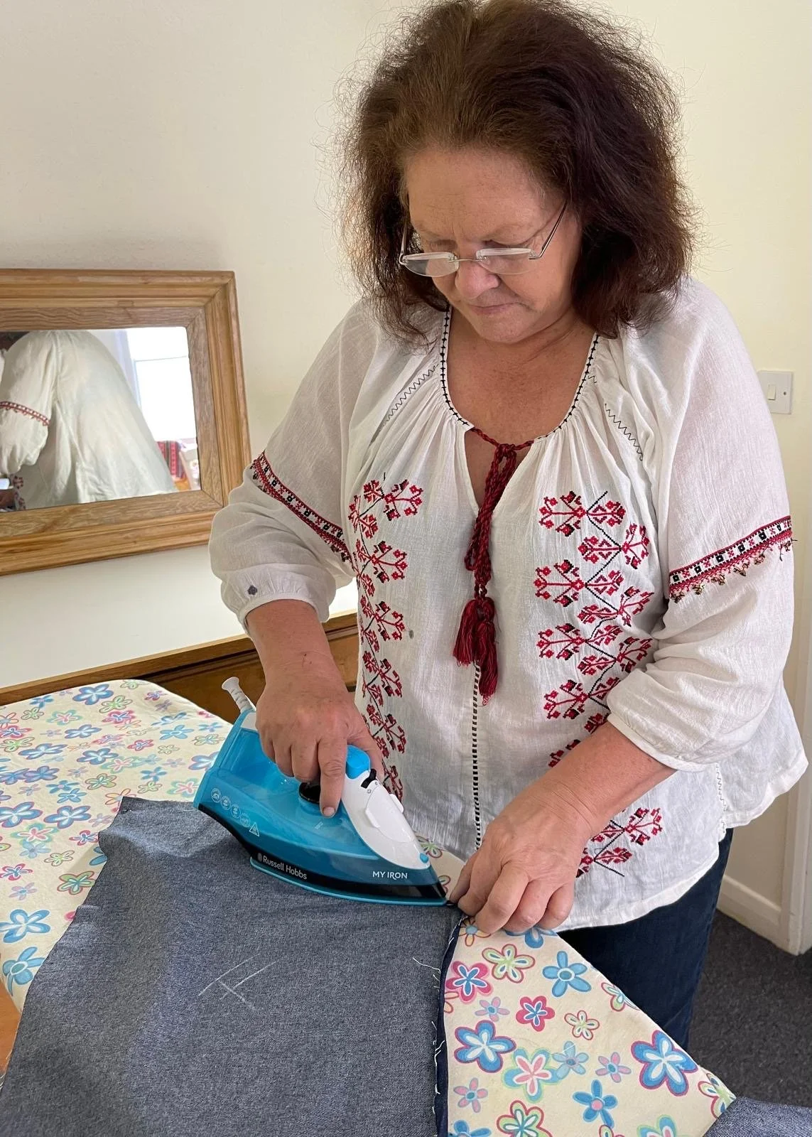 A woman with glasses wearing a white embroidered blouse is ironing a piece of dark fabric on a table with a colorful floral tablecloth. A mirror on the wall reflects another person in the background.