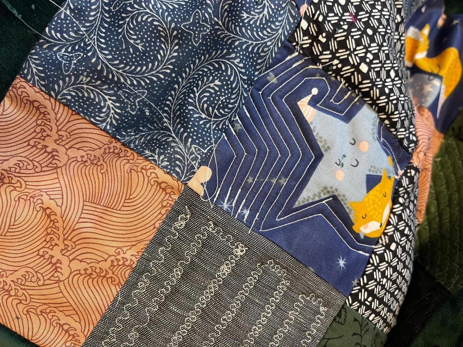 A patchwork quilt with various patterned fabrics, including designs of foxes, stars, geometric shapes, and floral motifs in colors like navy blue, peach, black, and green.