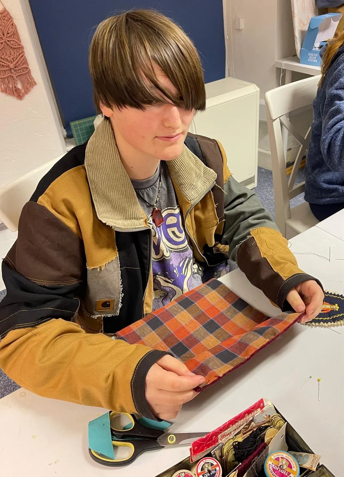 A young person with shoulder-length brown hair and fair skin sitting at a table examining a piece of fabric with a red, orange, and blue plaid pattern. They are wearing a tan and black jacket over a graphic T-shirt and a necklace with a red pendant. 