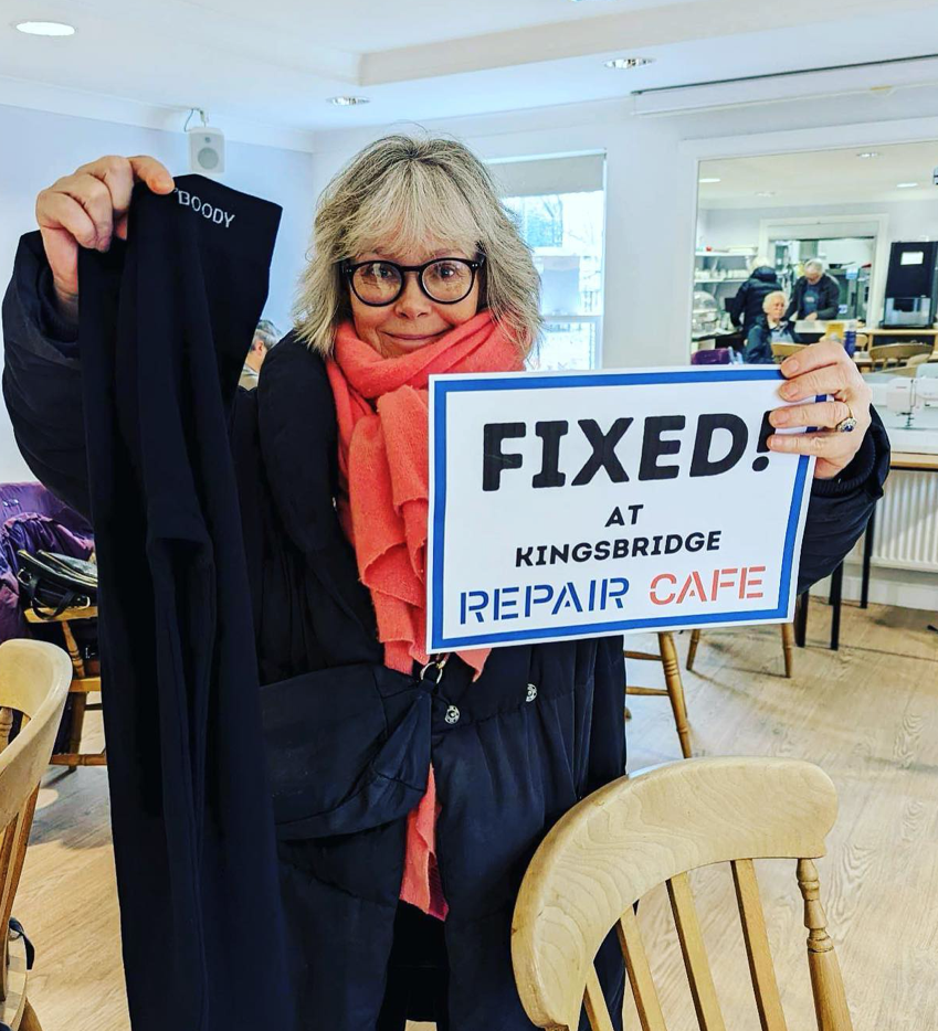 A woman with glasses and gray hair holding a sign that says 'FIXED! at Kingsbridge Repair Cafe.' She is also holding a piece of clothing. The cafe interior has light-colored walls, large windows, and a kitchen area with people working in the backgrou